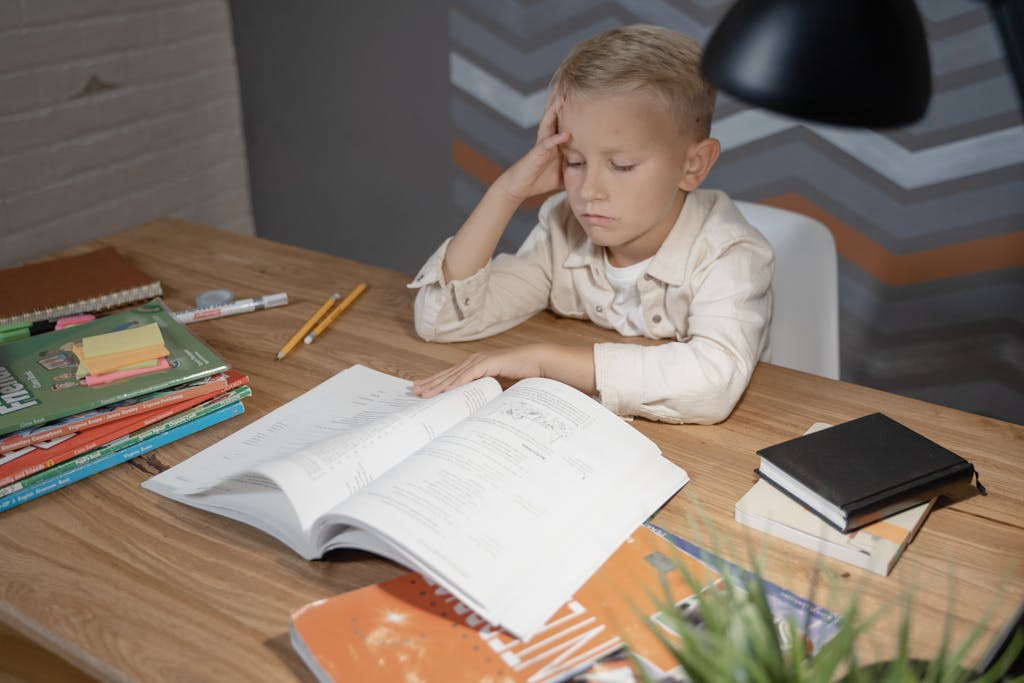 A young boy appears deep in thought while studying at his desk filled with books and notebooks.