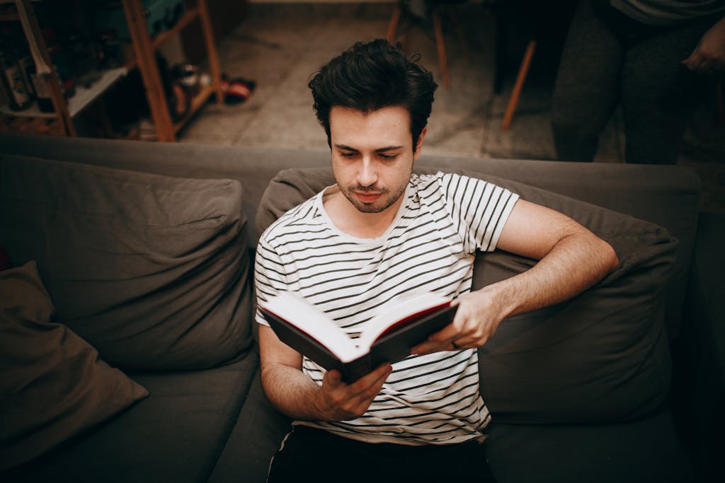 A young man relaxing on a sofa while reading a book in a cozy room.