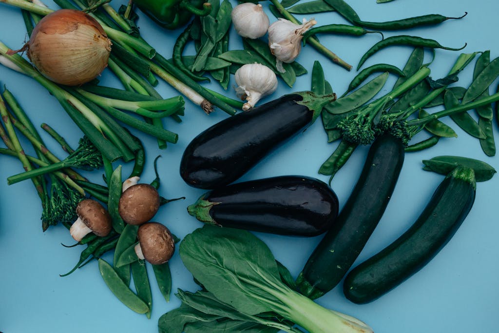 An overhead view of assorted fresh vegetables including eggplants, onions, and greens on a blue background.