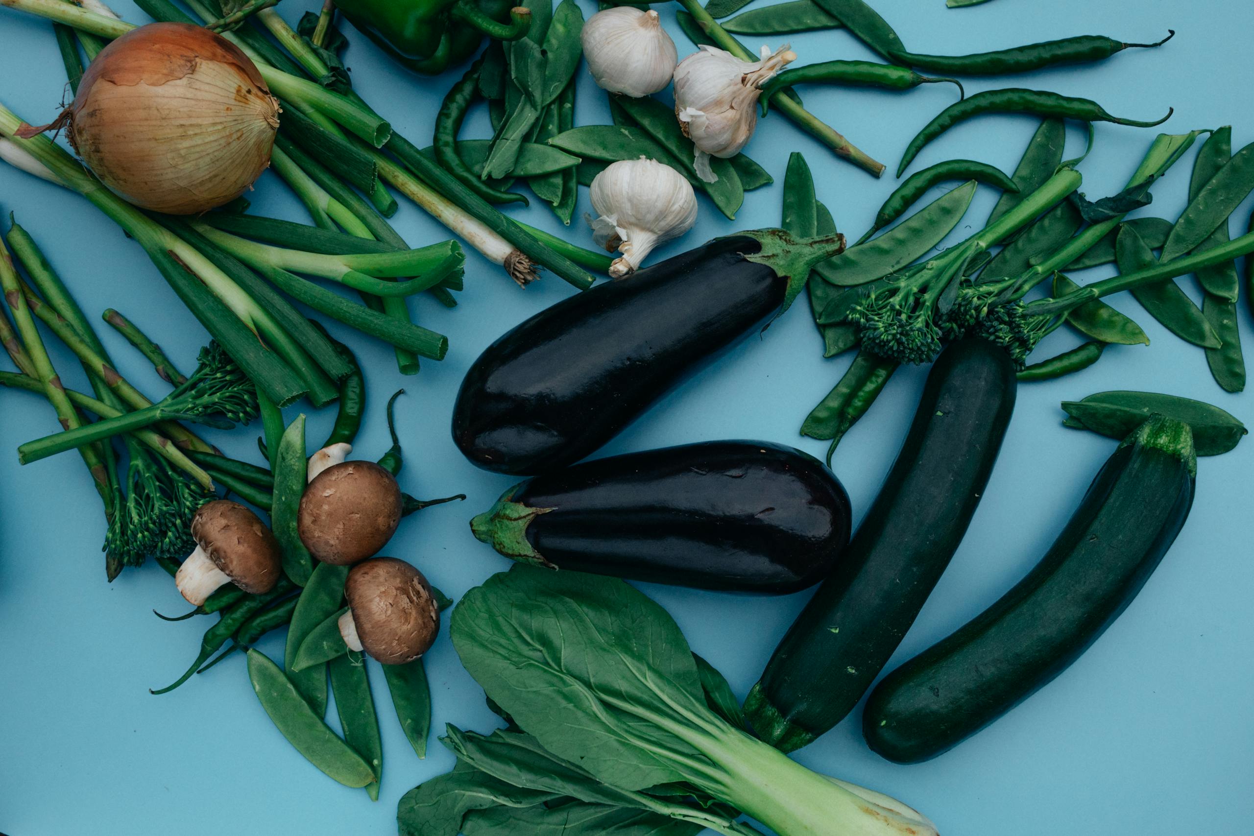 An overhead view of assorted fresh vegetables including eggplants, onions, and greens on a blue background.