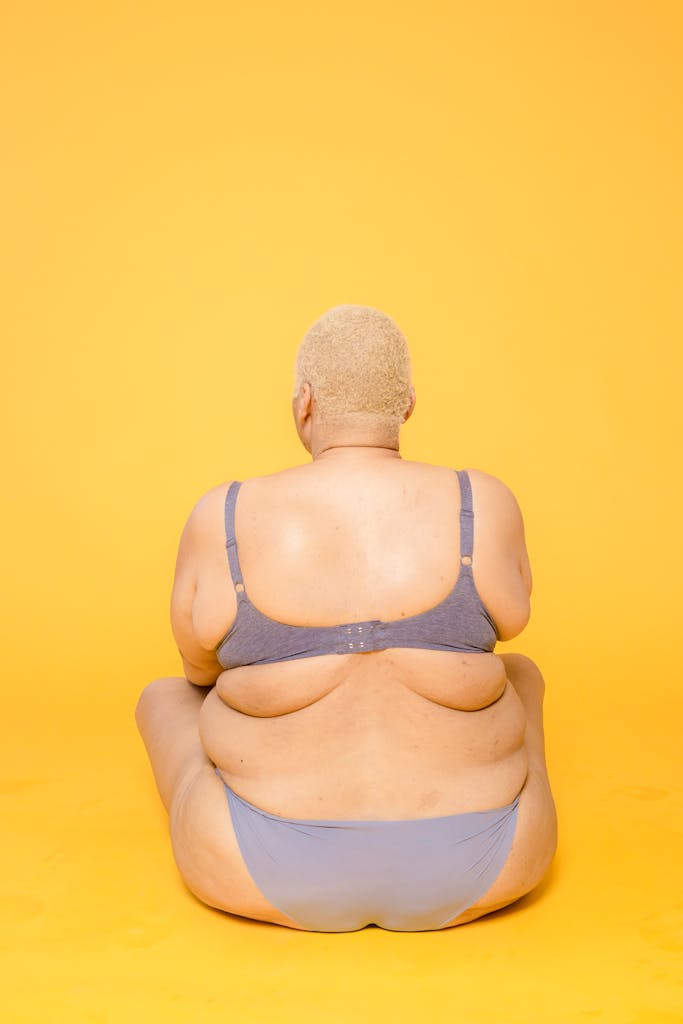 Back view of anonymous plump female model with short hair wearing lingerie sitting on floor on yellow background in studio