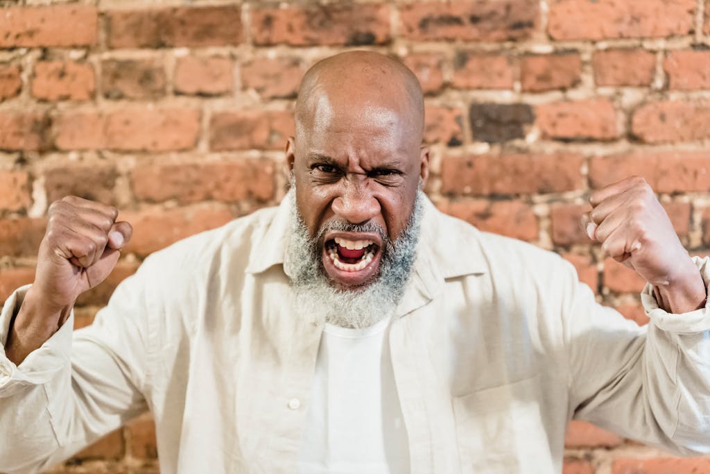 Bald man showcasing strong emotions with fists clenched in front of a brick wall.