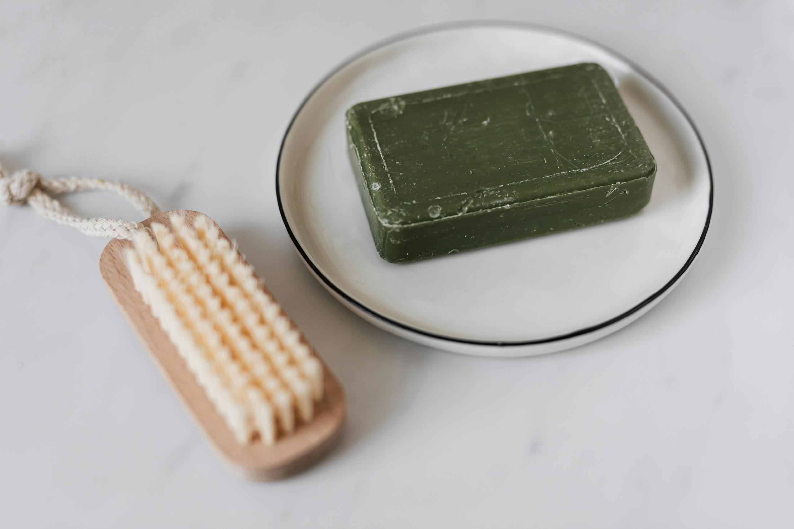 Close-up of a green soap bar on a dish with a wooden brush on a marble surface.