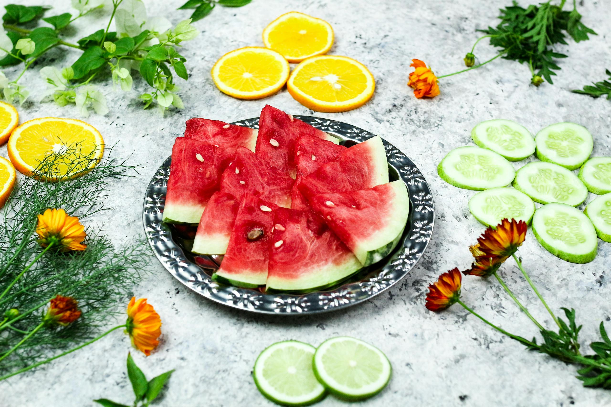 Colorful assortment of watermelon, citrus, and cucumbers with flowers on marble.