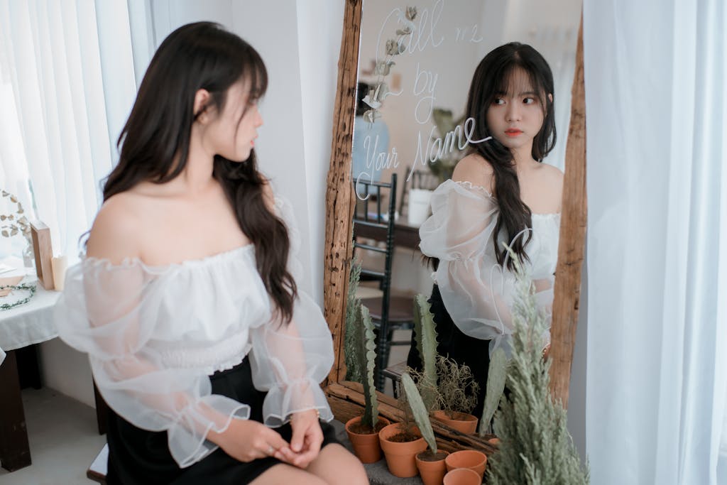 Elegant woman in white blouse looking in mirror indoors.