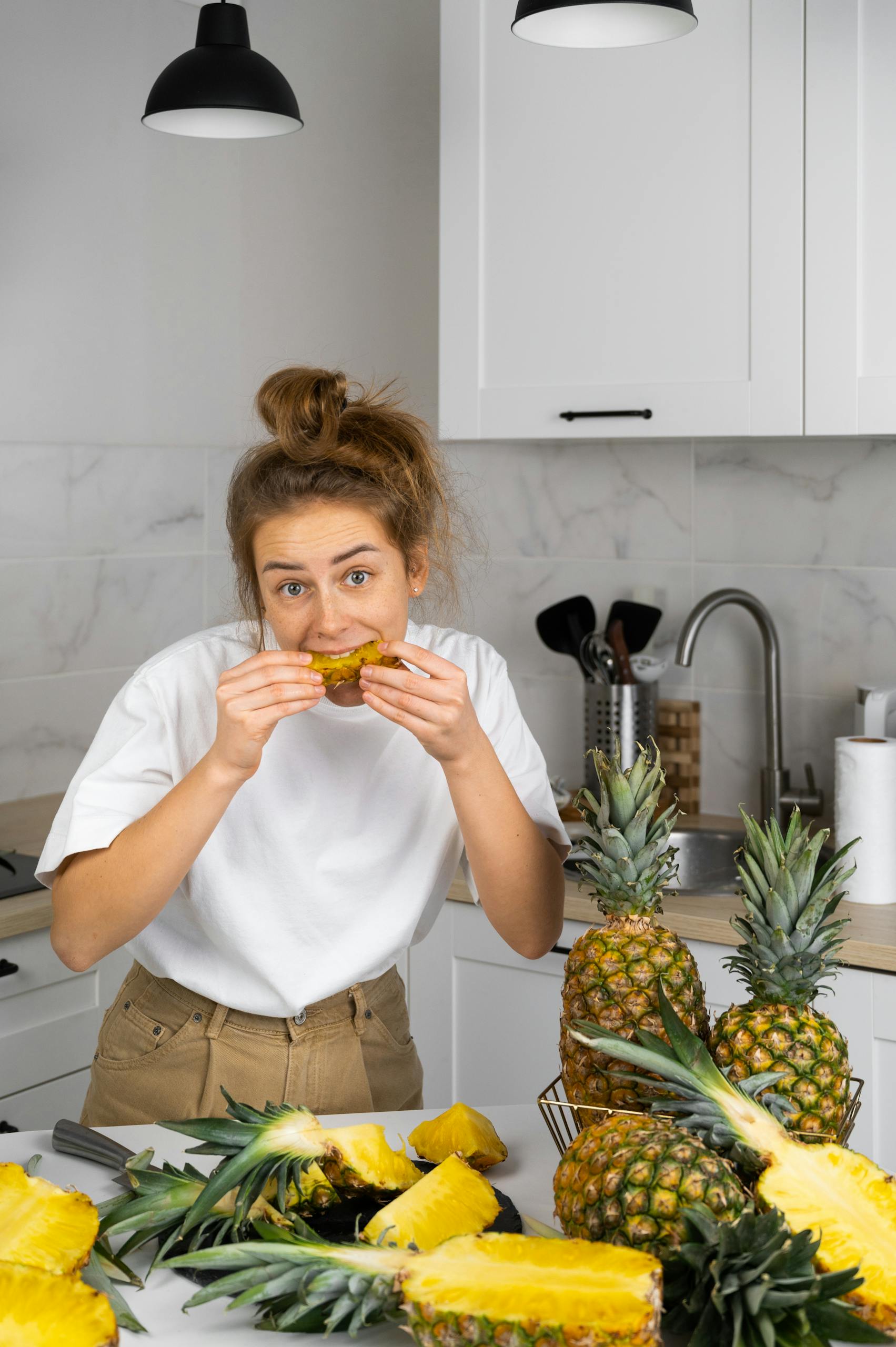 Female in white shirt standing at table while eating juicy pineapple in kitchen and looking at camera