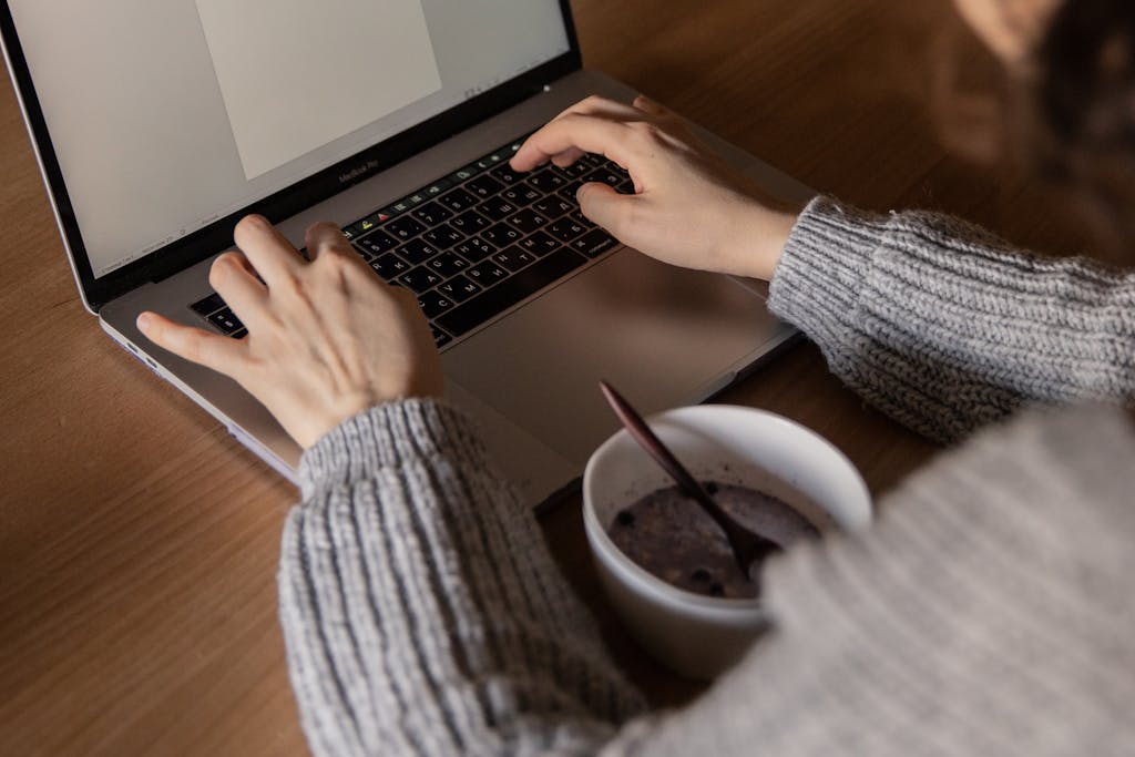 From above unrecognizable woman in gray sweater sitting at wooden table with food in bowl and creating document while working on remote project at home
