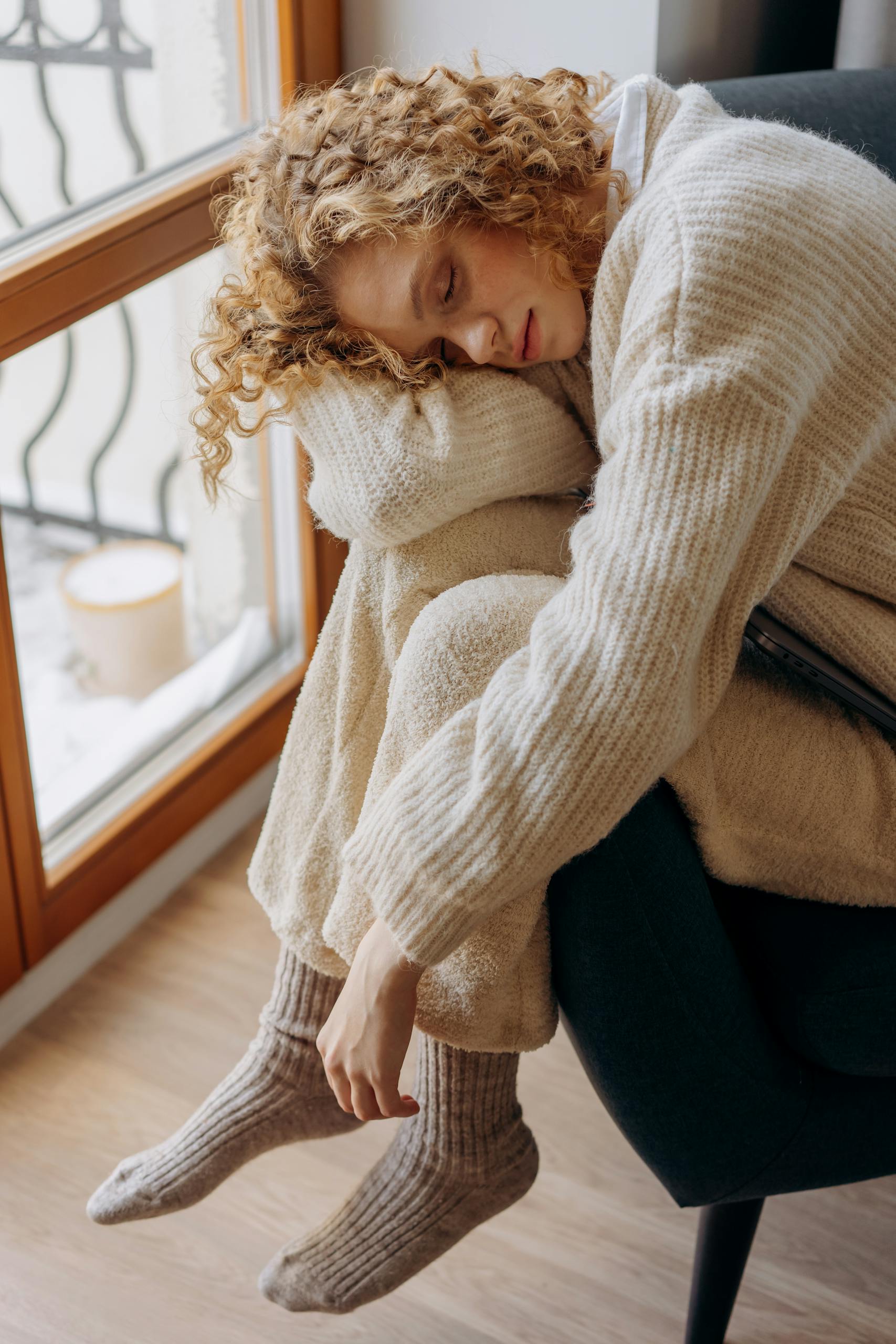 Peaceful woman in a cozy knitted sweater resting by the window on a sunny day.
