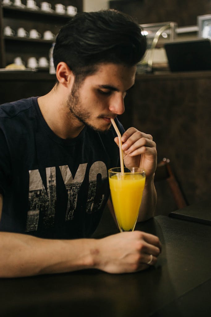 Side profile of a young man with dark hair sipping orange juice through a straw indoors.
