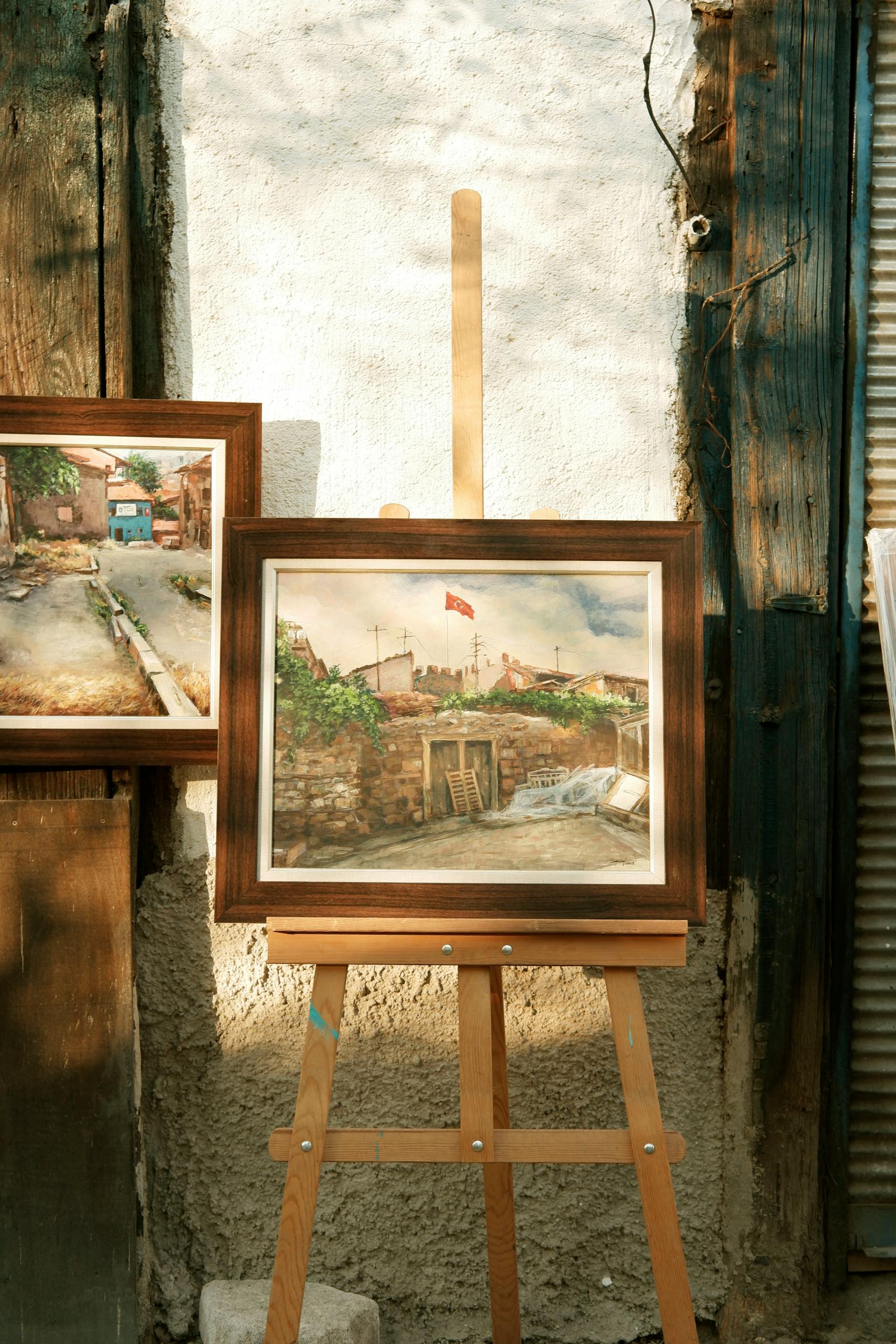Two framed landscape paintings displayed on an easel against a rustic wall.