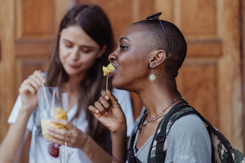 Two women savoring fruit skewers outdoors, capturing a moment of enjoyment and hunger satisfaction.