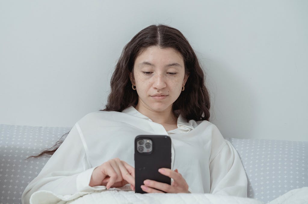 Young female with long dark wavy hair in white nightwear lying on comfortable bed under white blanket and browsing mobile phone in morning time at home