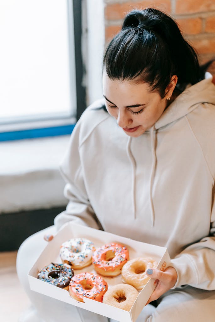 Young plump female in casual clothes taking box with assorted yummy fresh donuts in light room