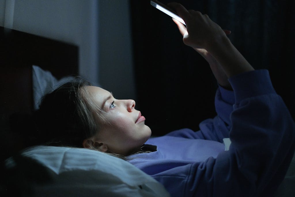Young woman lying in bed using a smartphone at night, illuminated by screen light.