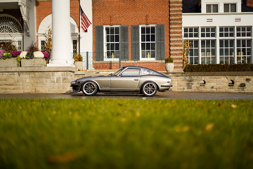 A classic silver sports car parked in front of a luxurious mansion with an American flag displayed.