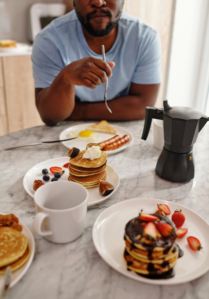 A close-up of a man's breakfast with pancakes, fruit, and a moka pot on a marble table.