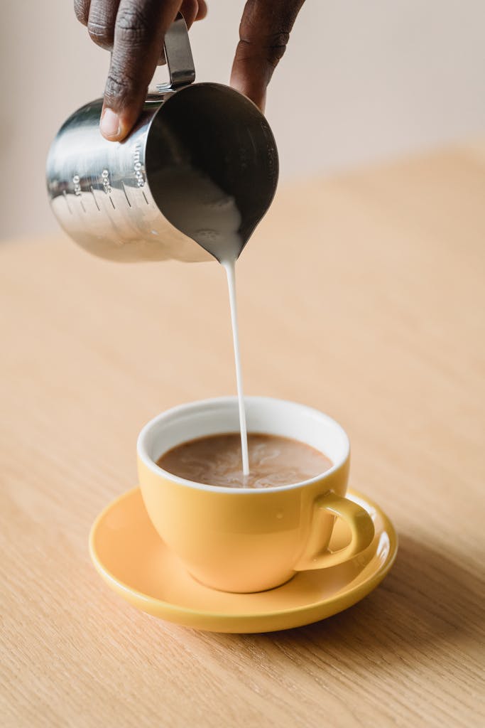 A close-up shot of milk being poured into a yellow coffee cup on a wooden table.