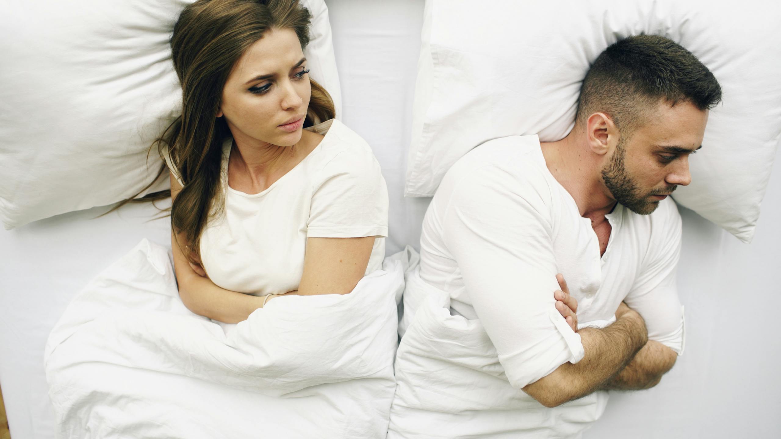A couple in bed, facing away with tense expressions, suggesting conflict.