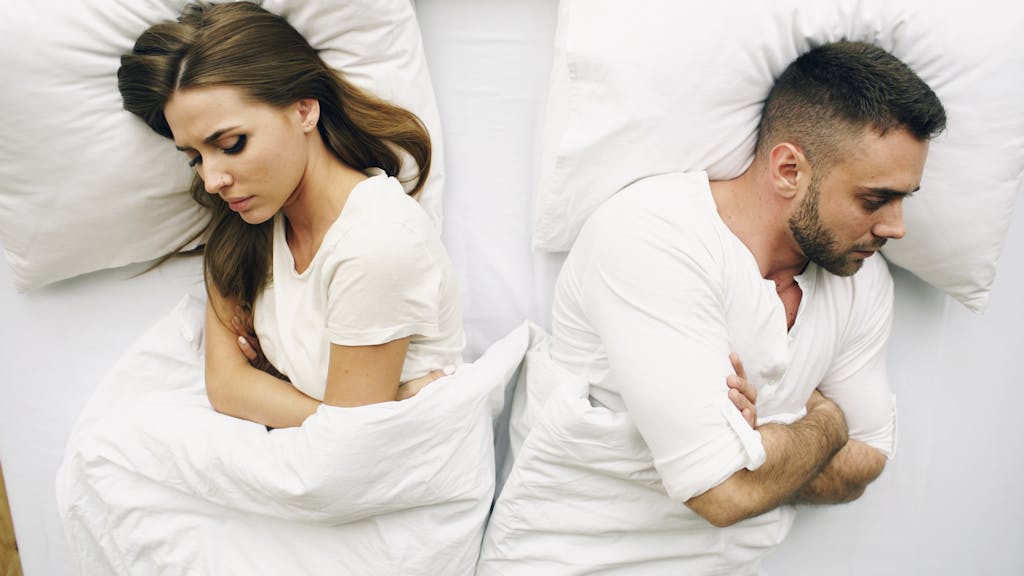 A couple lying in bed facing away, expressions of discontent.
