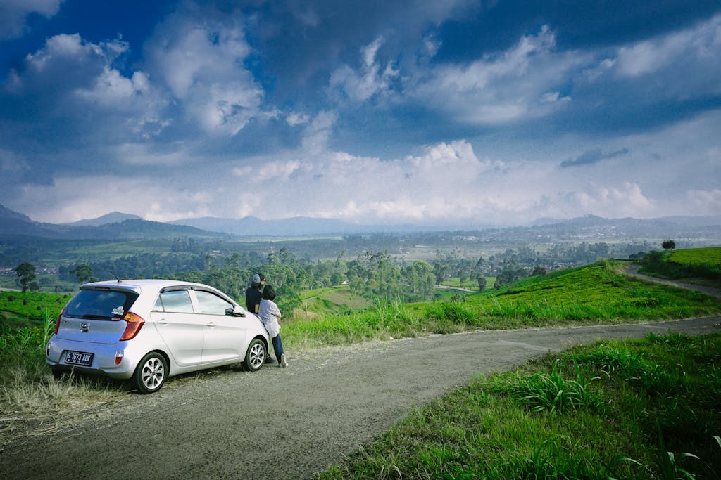 A couple stands by their car on a road overlooking lush green fields and mountains under a cloudy sky.