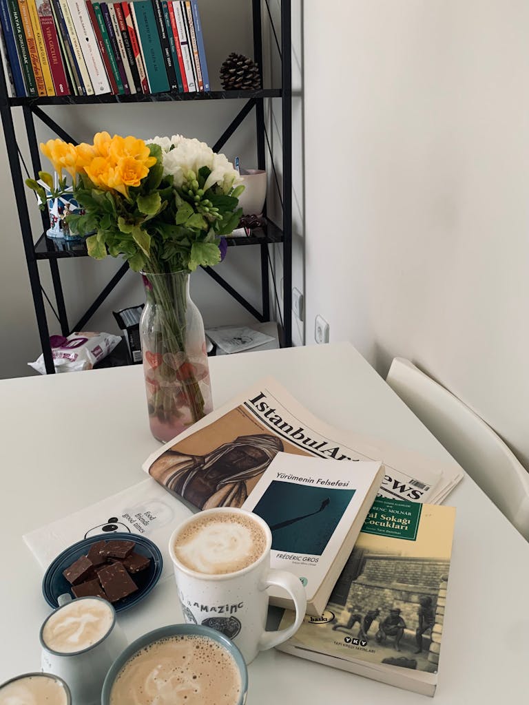 A cozy indoor setup with books, cappuccino, chocolates, and flowers on a table.