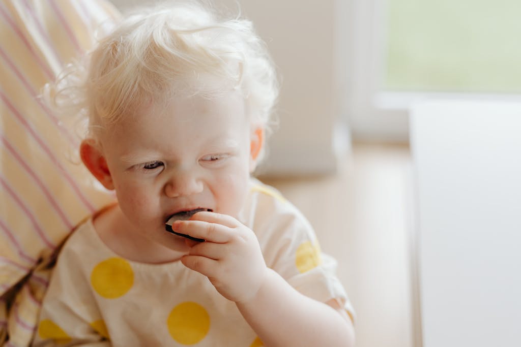 A cute toddler with blonde hair enjoys a snack indoors, capturing innocence and joy.