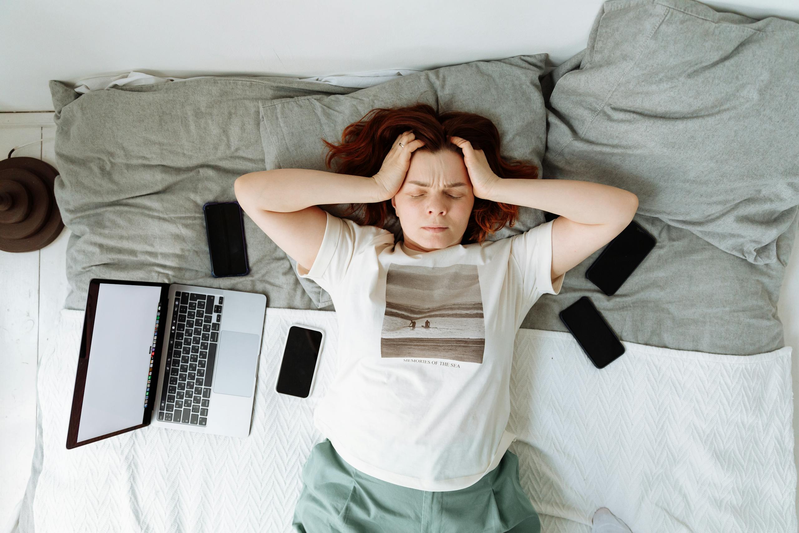 A distressed woman lying in bed surrounded by digital devices, showing signs of stress.