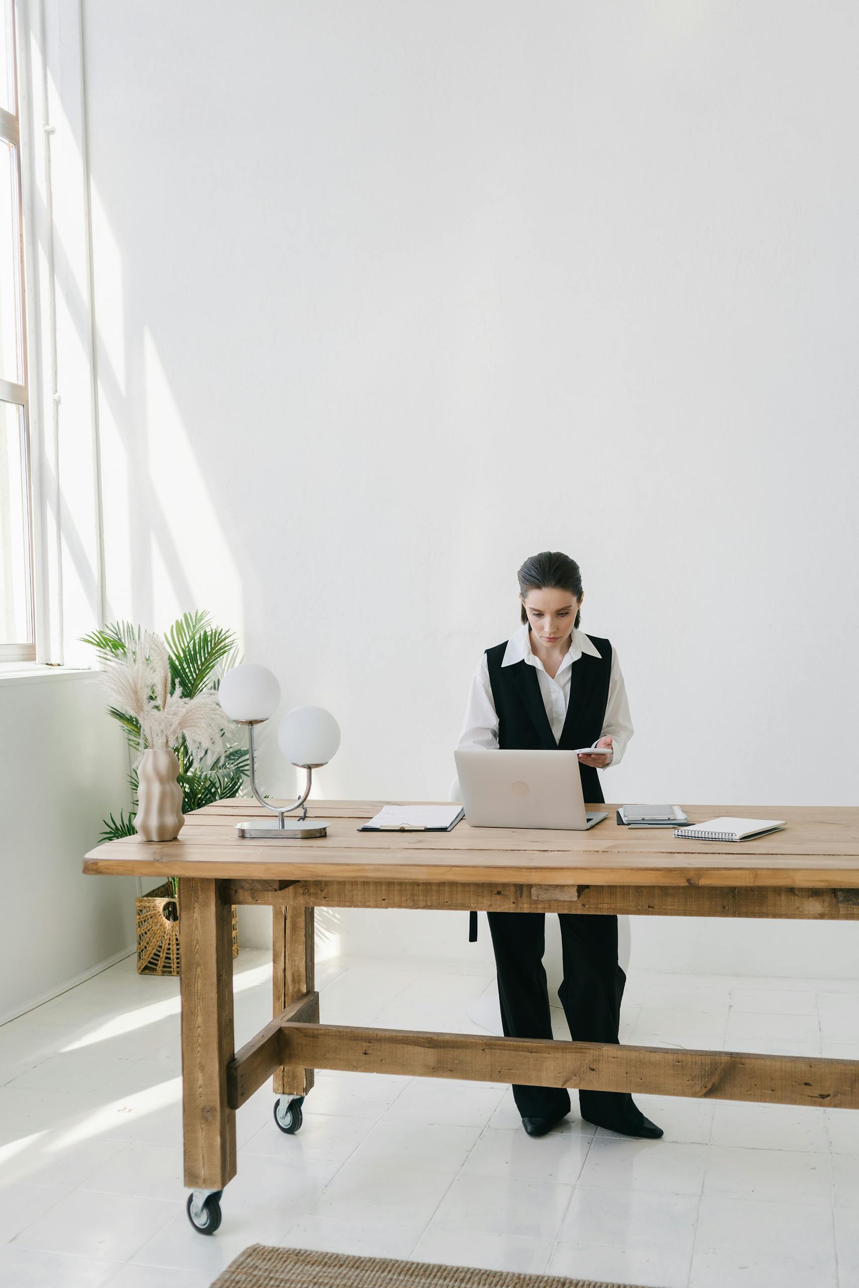 A focused woman stands at a wooden desk, working on her laptop in a well-lit office space.