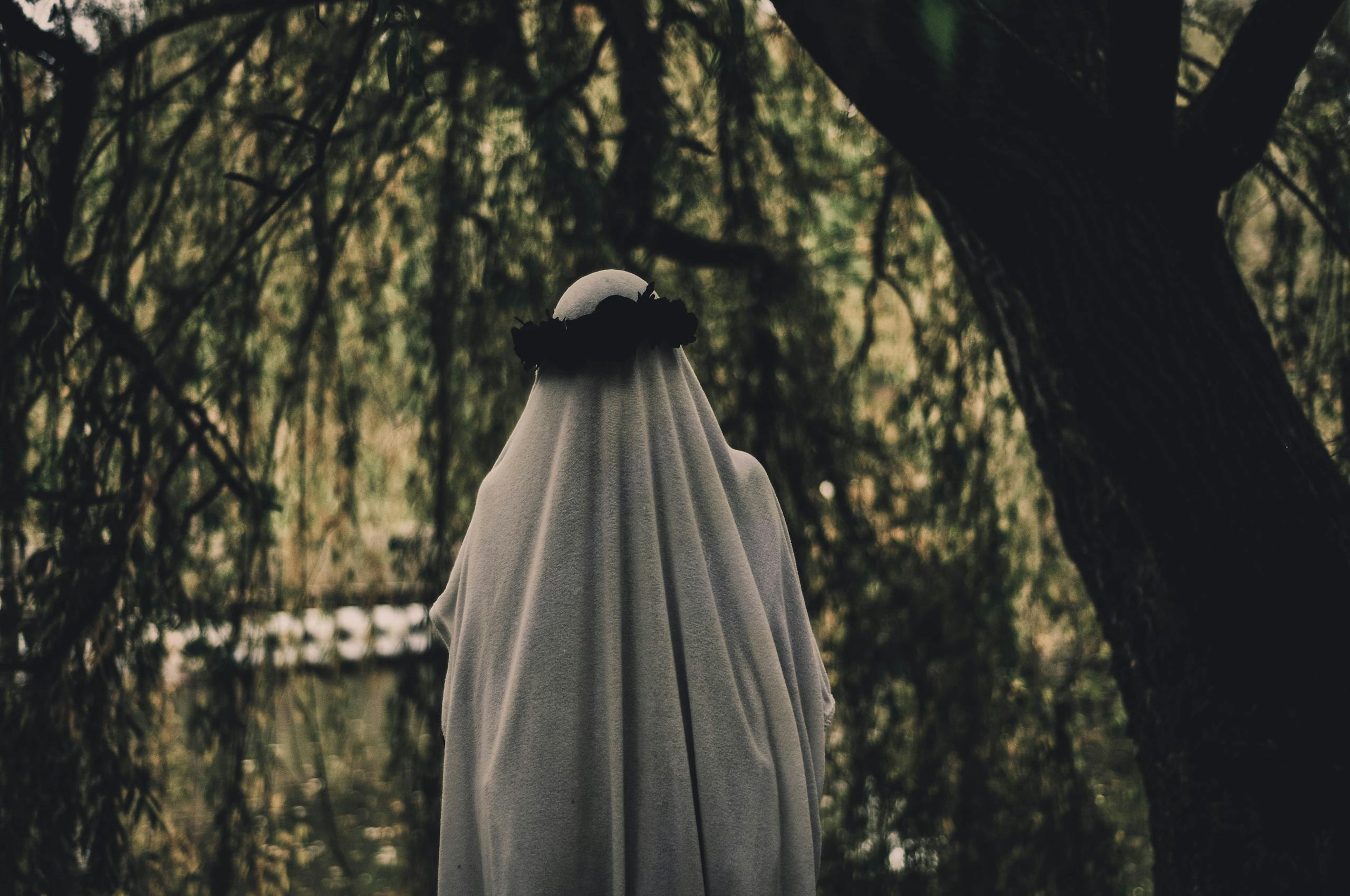 A ghostly figure draped in white stands in a dimly lit park surrounded by trees.