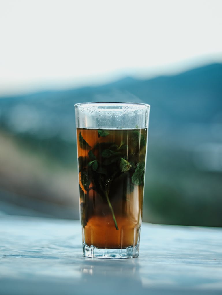 A glass of refreshing Moroccan mint tea with fresh herbs, set against a serene mountain backdrop, captured outdoors.