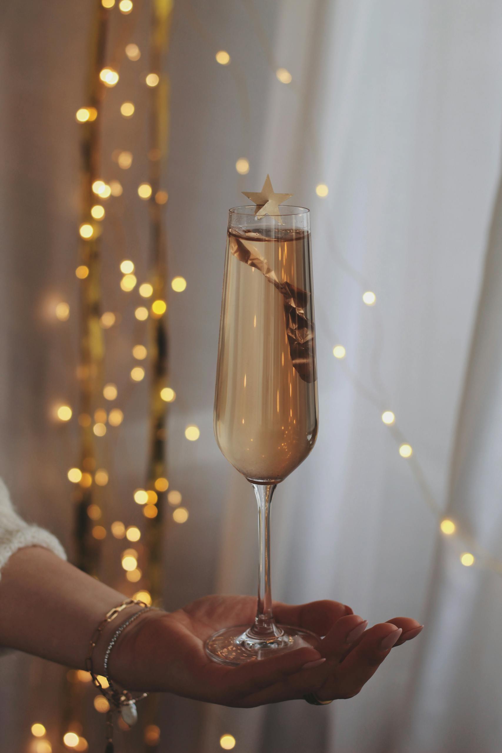 A hand holding a champagne glass adorned with a star topper, set against twinkling fairy lights.