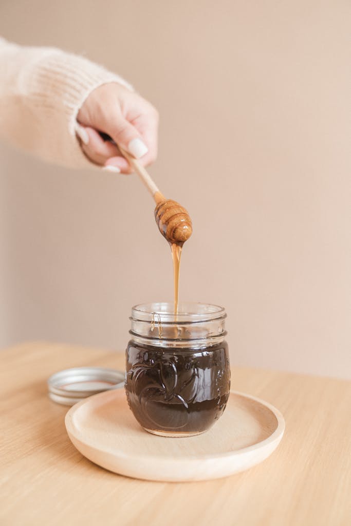 A hand holding a honey dipper drizzles honey into a jar on a wooden plate indoors.