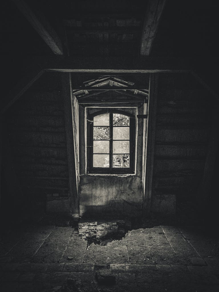 A haunting monochrome image of an abandoned attic, featuring a broken window and eerie shadows.