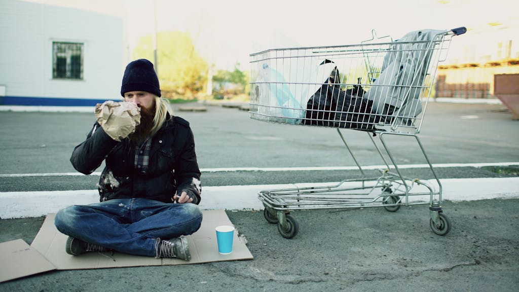 A homeless man sits on cardboard drinking from a bag beside a shopping cart outdoors.