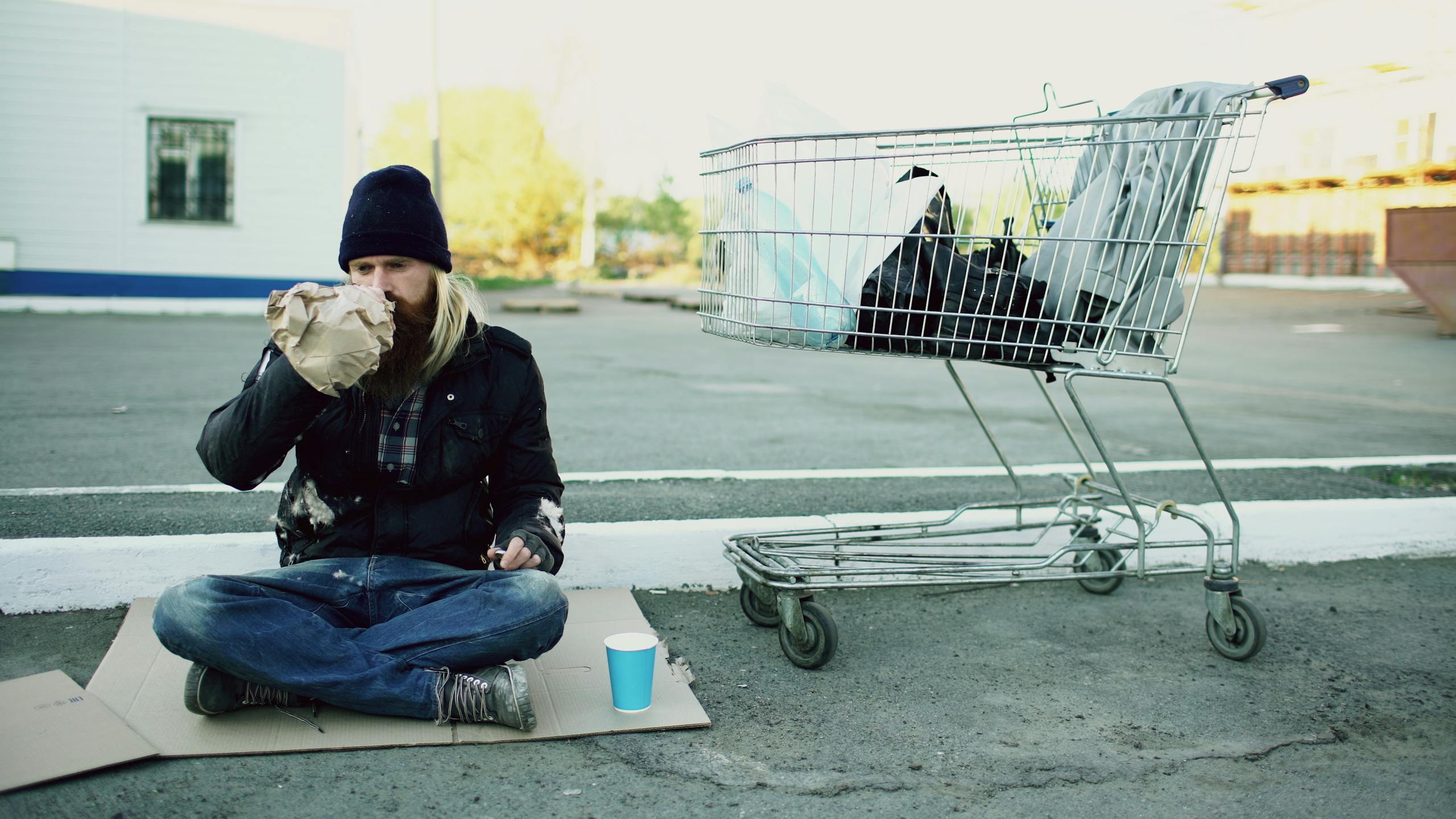 A homeless man sits on cardboard drinking from a bag beside a shopping cart outdoors.