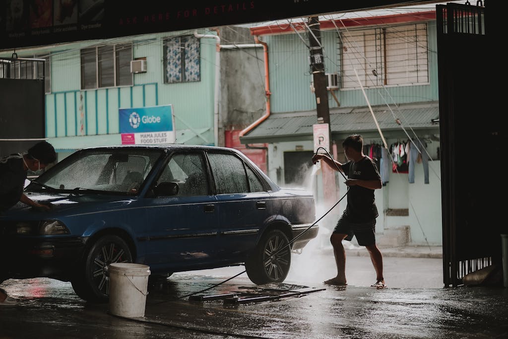 A man cleaning a blue car at an outdoor carwash with a hose, urban setting.