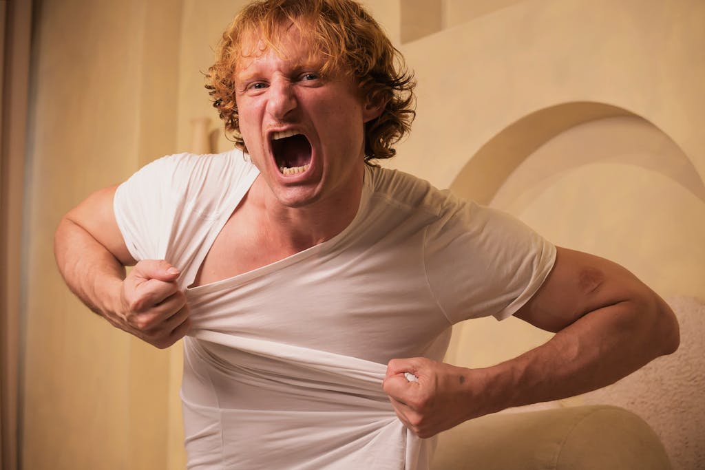 A man exhibiting anger and frustration by tearing his shirt indoors, with an intense expression.