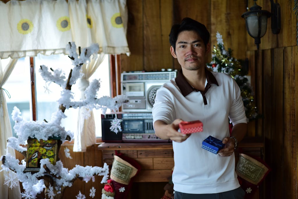 A man in a cozy room decorated for Christmas offering a red gift box.