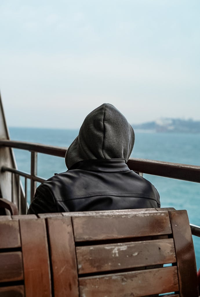 A man in a hoodie sits on a wooden bench beside the sea, gazing thoughtfully at the horizon.
