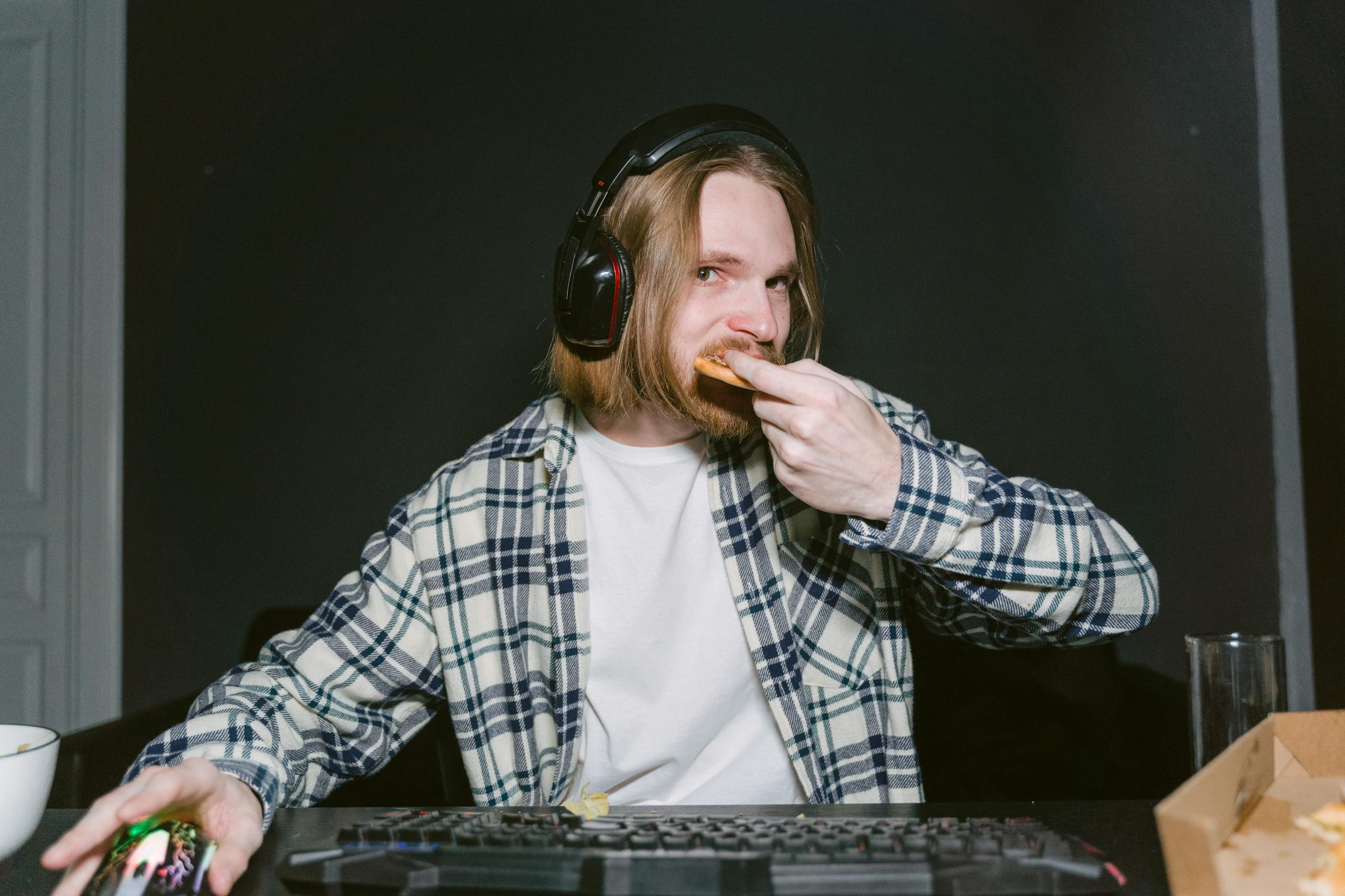 A man wearing headphones eats pizza while gaming on a computer, focused and engaged.