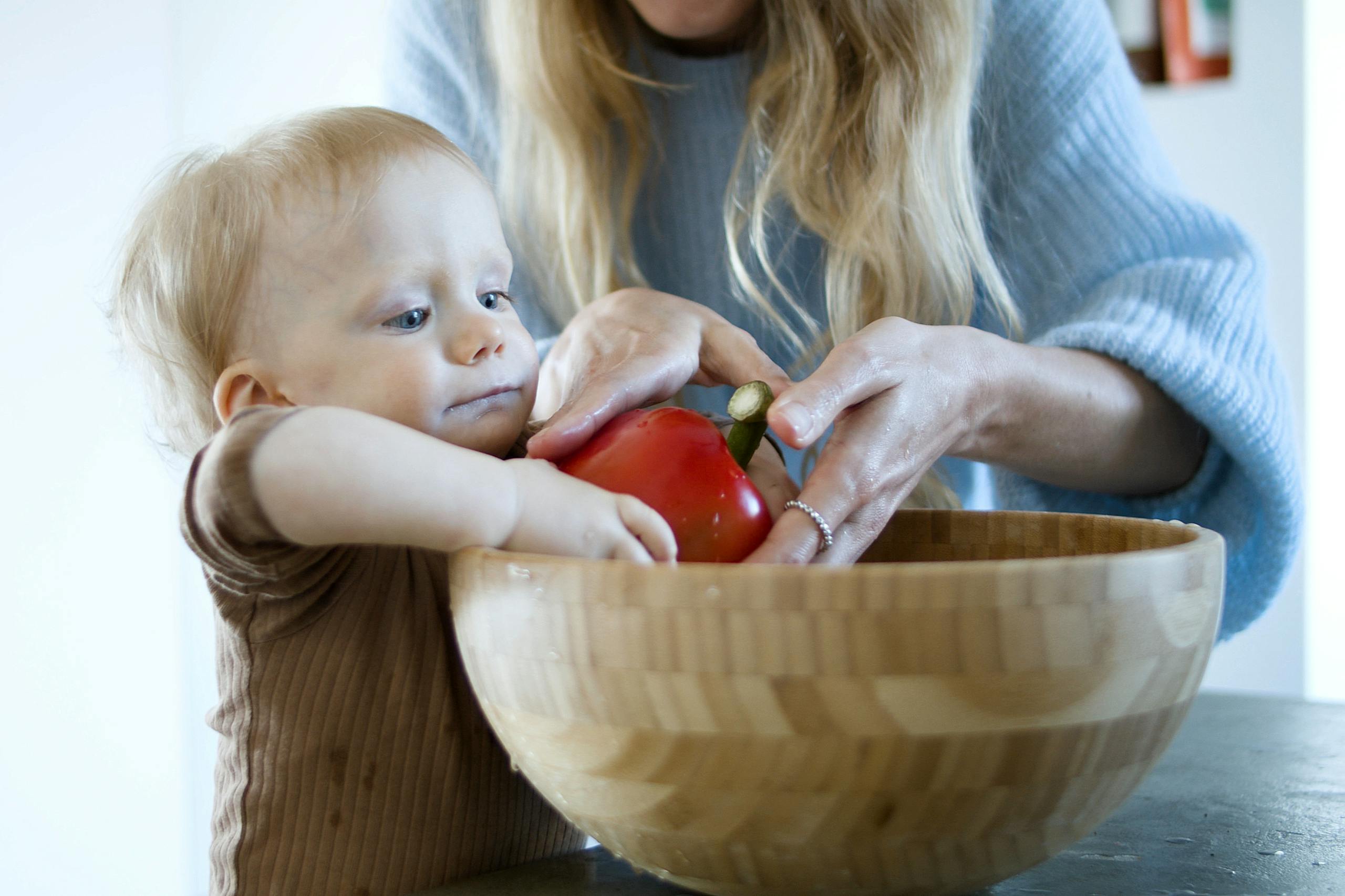 A parent helps a young child wash a red bell pepper in a wooden bowl, promoting healthy eating habits.