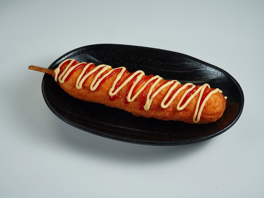 A perfectly cooked corndog topped with ketchup and mayonnaise on a black plate.
