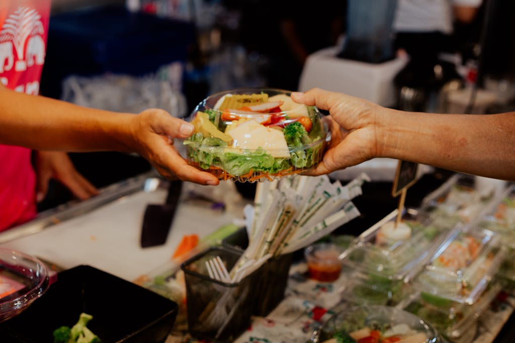 A person hands over a fresh salad in a plastic container at a food stall.