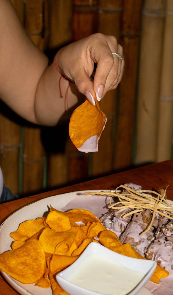 A person holds a sweet potato chip above a plate with sauce, ready to dip.