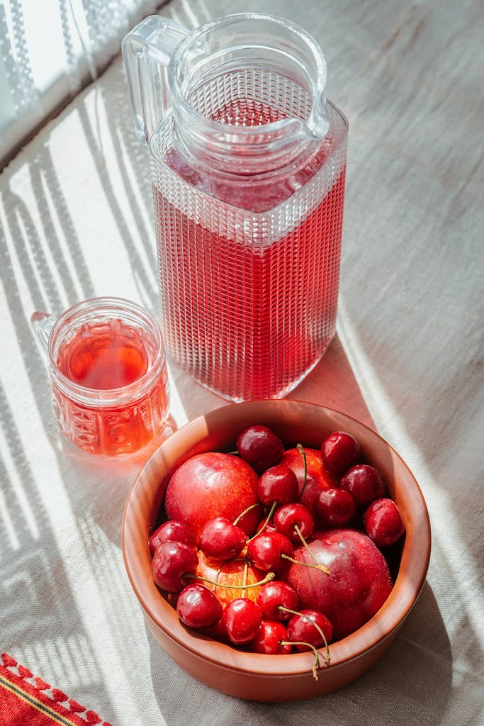 A rustic still life featuring cherries and nectarines in a bowl with red juice in a glass pitcher.