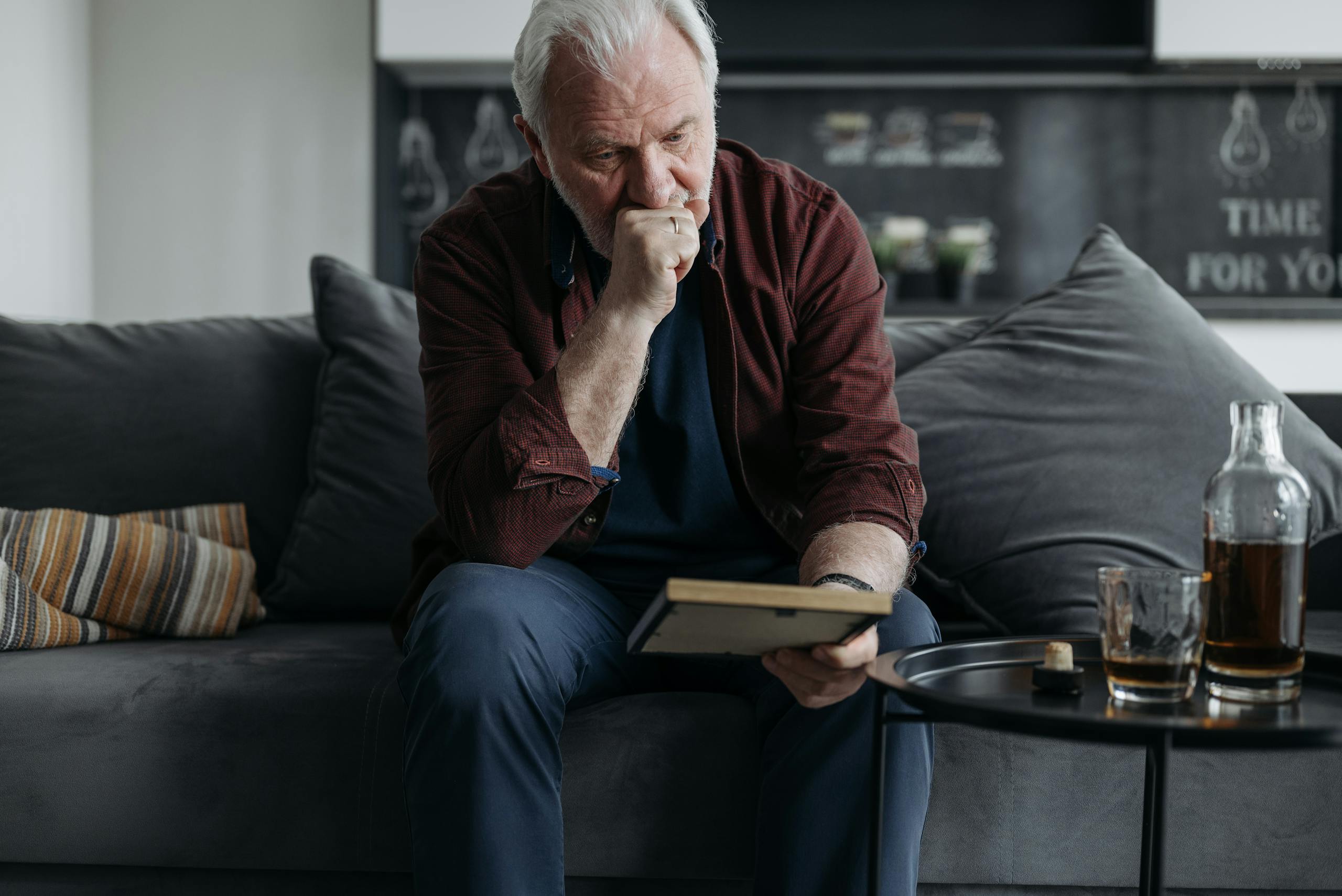 A senior adult man sitting on a couch, lost in thought while reading a book indoors.