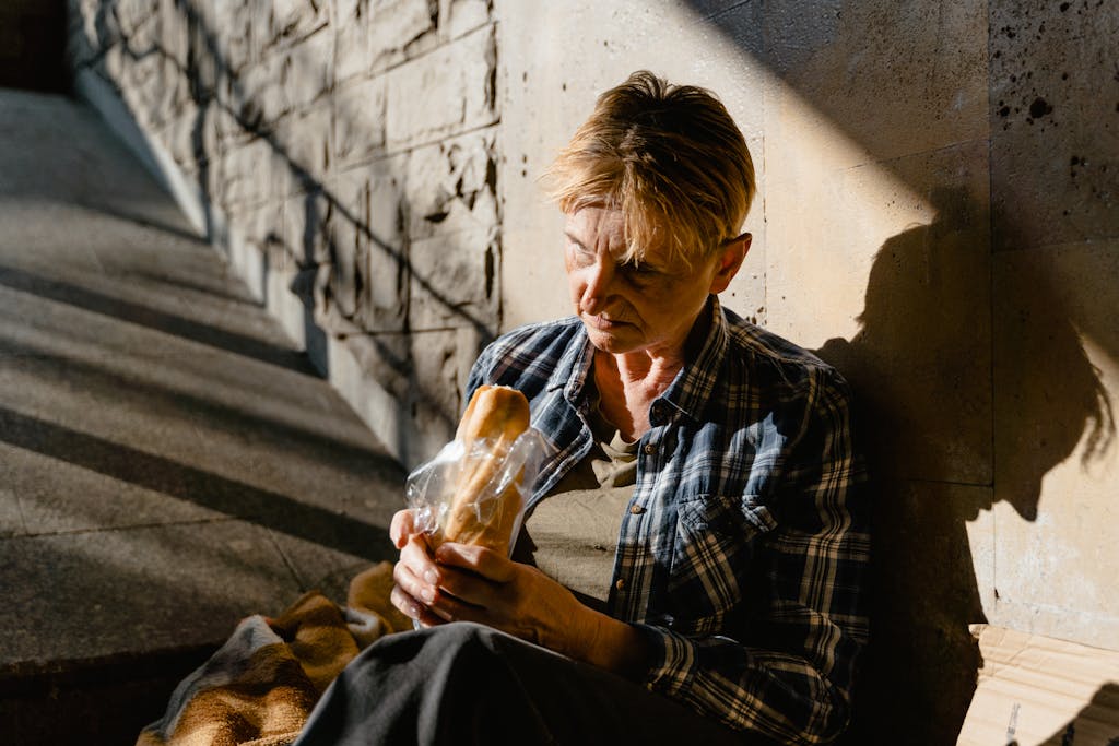 A senior woman holding and eating bread while seated against a sunlit wall, casting a thoughtful shadow.