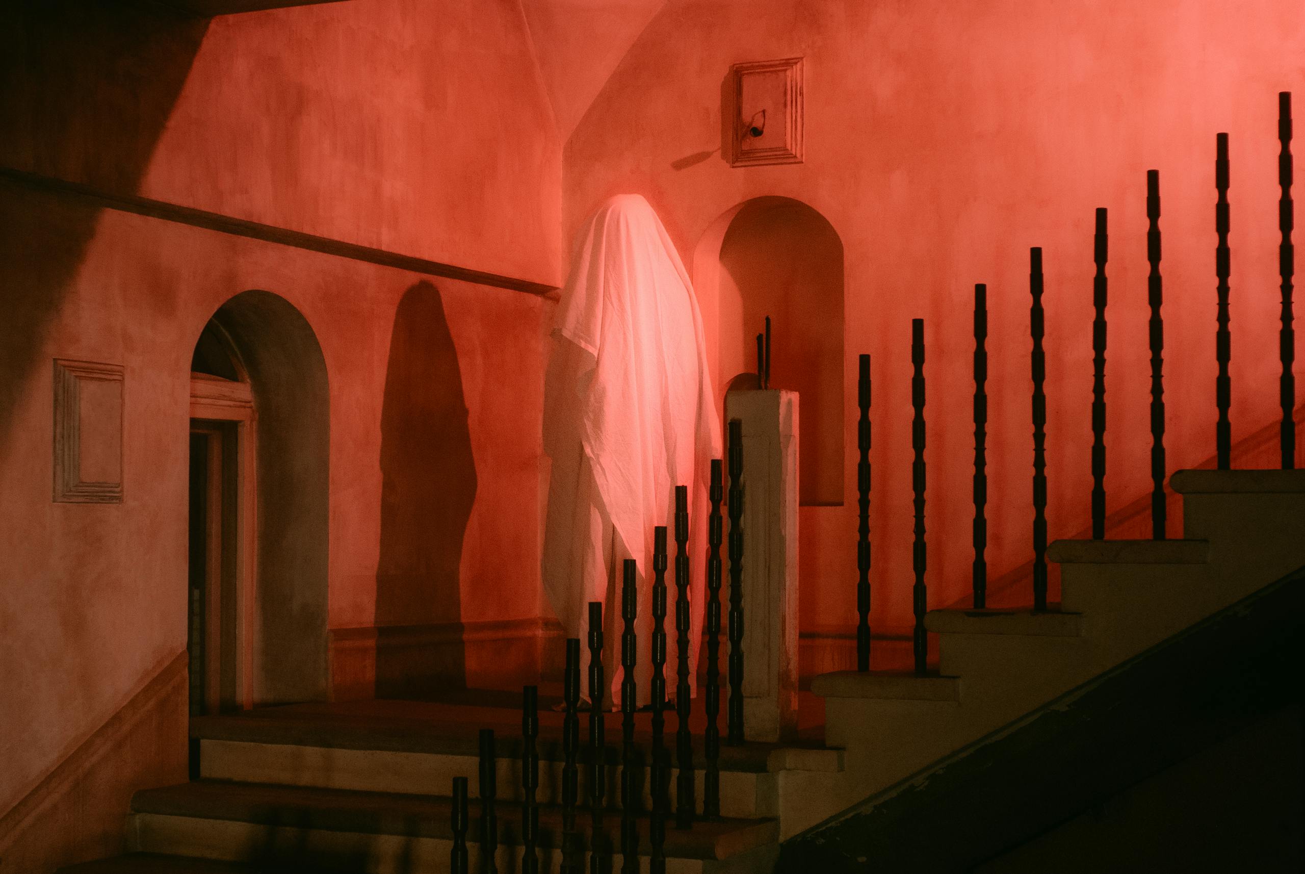 A spooky scene featuring a mysterious ghostly figure draped in white on a dimly lit, red-hued staircase.