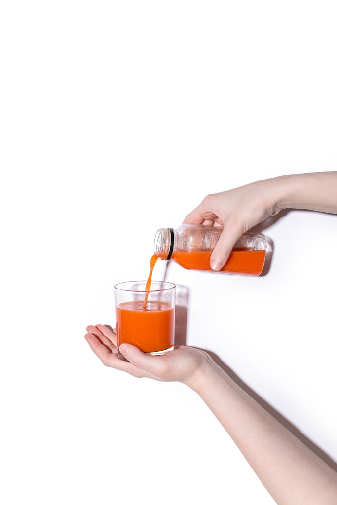 A vibrant carrot juice being poured into a glass from a bottle held by hands.