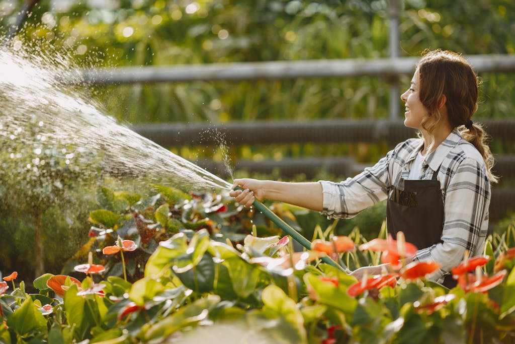 A woman in a plaid shirt and apron joyfully waters colorful flowers in a lush garden.